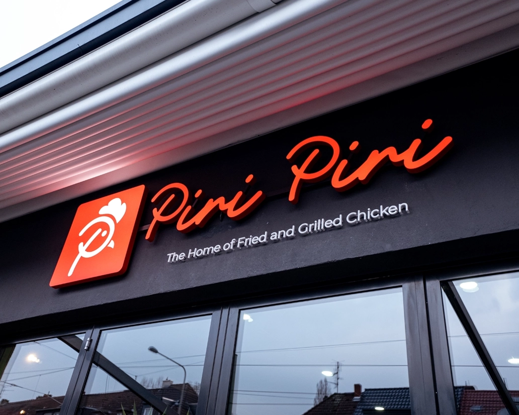 Modernes, beleuchtetes LED-Logo der Marke „Piri Piri – The Home of Fried and Grilled Chicken“ an einer schwarzen Gebäudefassade in Frankfurt – Branding-Projekt von FourMedia.